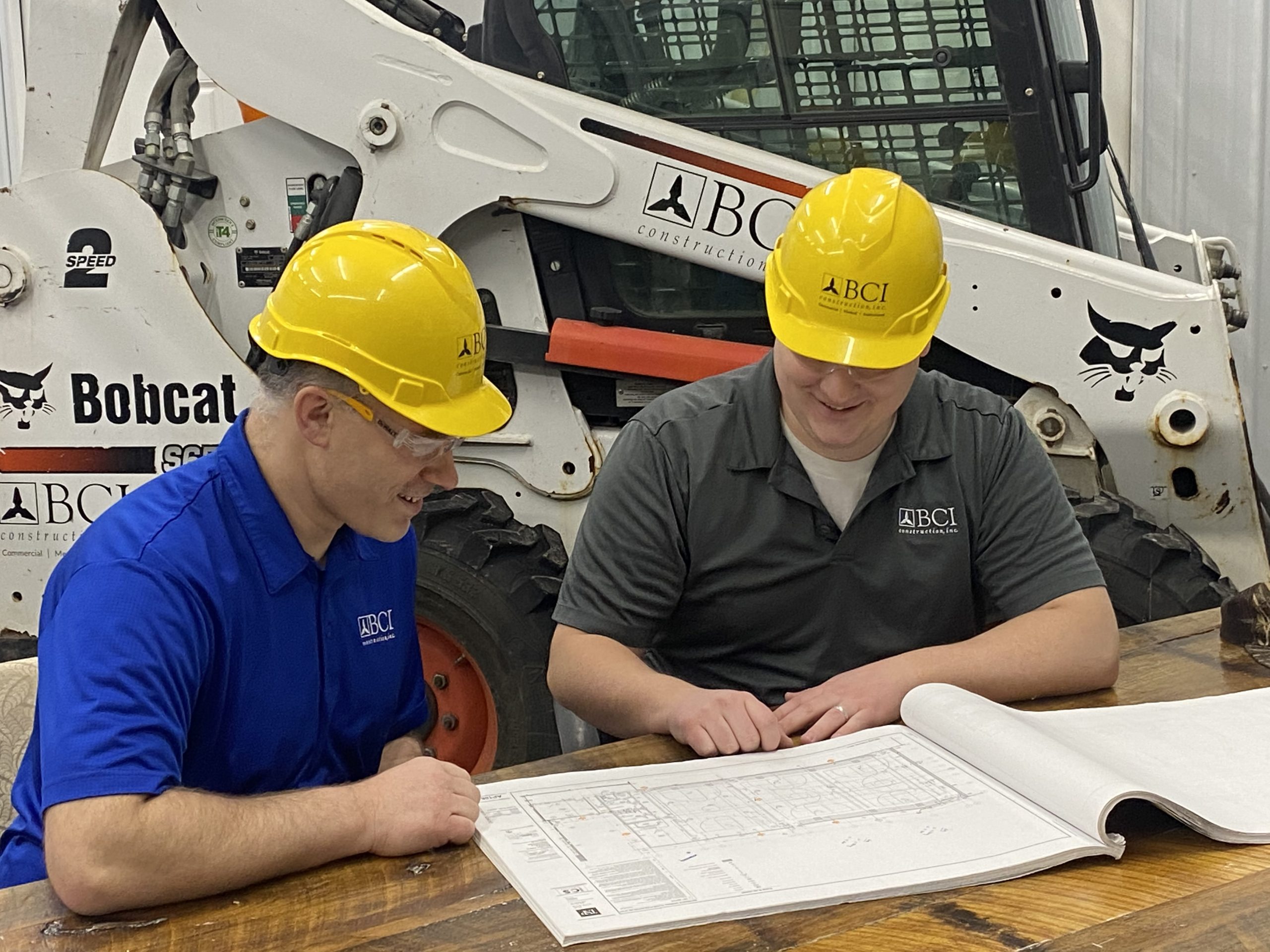 Two construction workers wearing hardhats sit at a table and discuss plans over blueprints with a bobcat in the background